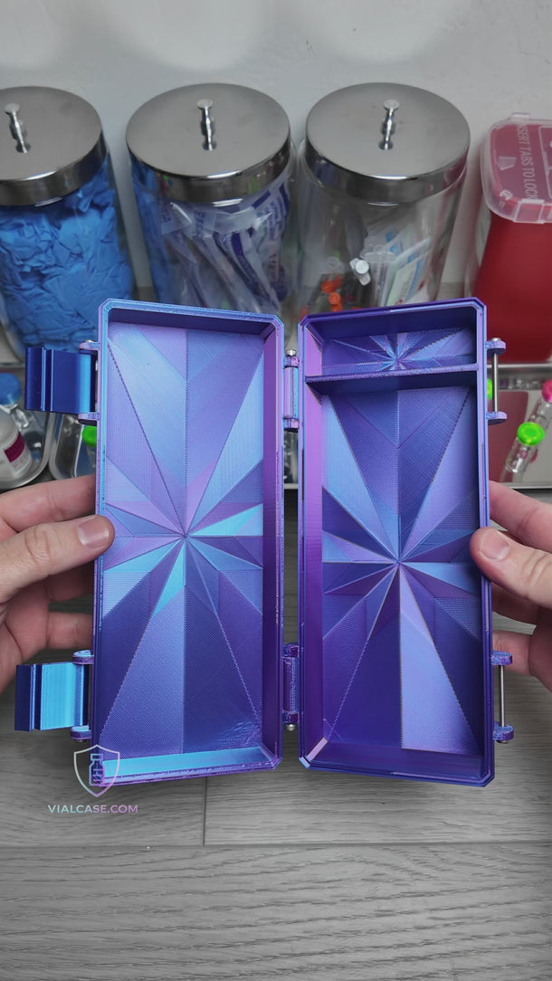 RGB metallic storage case with a mirror-like reflective finish opened on a tabletop, showing compartments holding prefilled syringes and alcohol prep pads. A hand places and adjusts items inside. In the background, trays of small vials with colorful caps and medical supplies are visible. The case has two latches and a structured interior for organized storage.